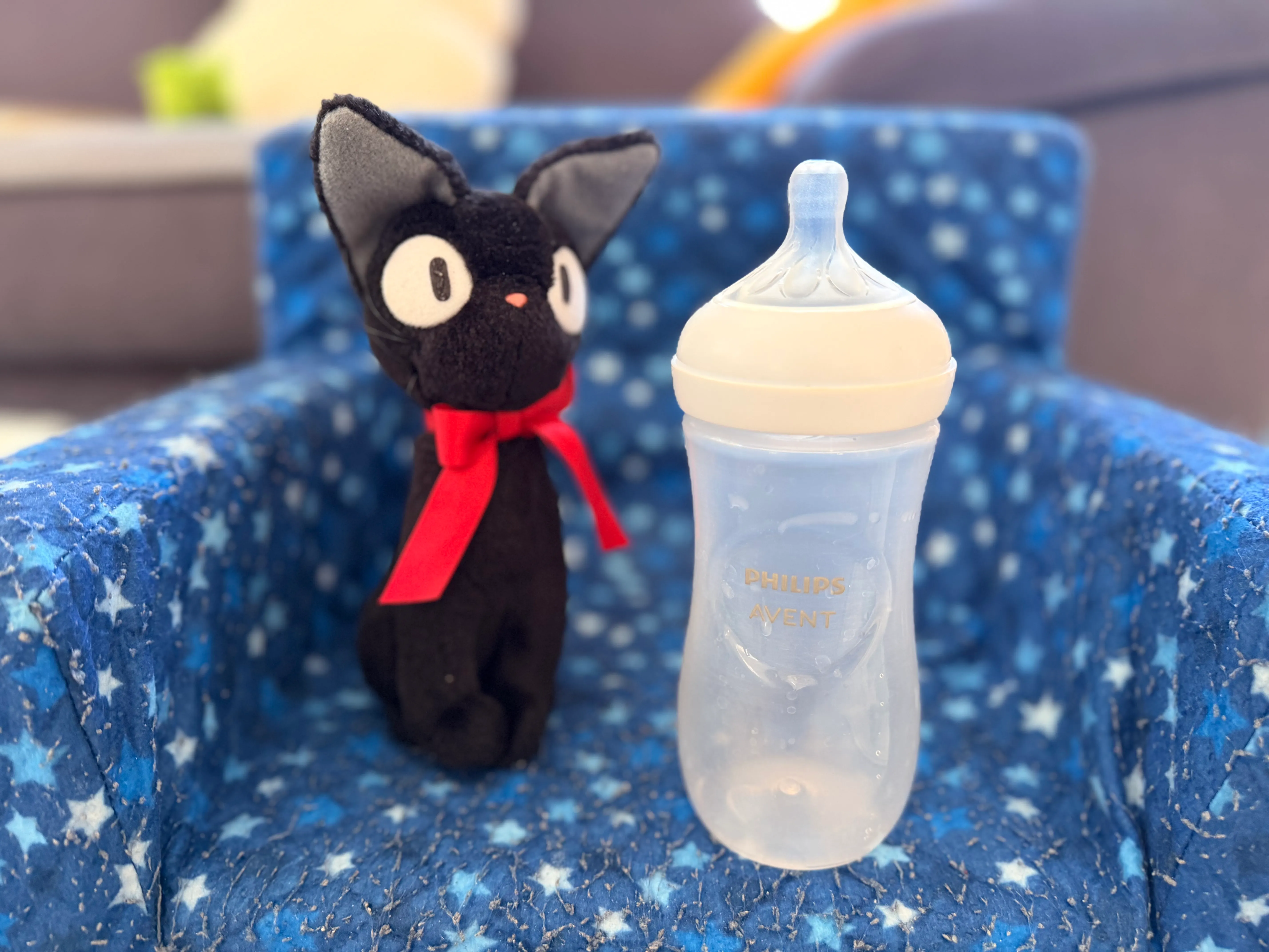 An empty baby bottle next to a small black cat plush toy, on a star-patterned blue armchair.